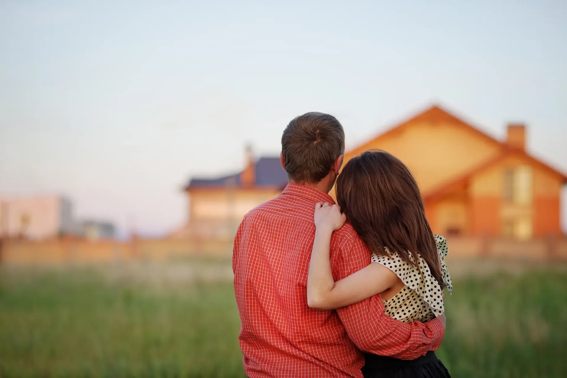 A couple faces away from camera and looks at a house in the distance.